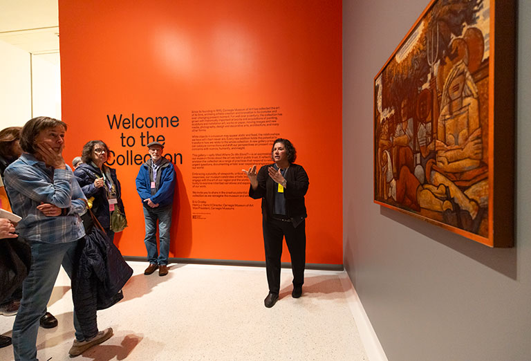 A museum guide engages a small group, highlighting an artwork on a wall next to an orange "Welcome to the Collection" display.