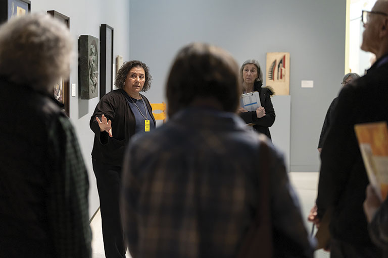 A group of visitors listens attentively as a guide shares insights in an art gallery, surrounded by various artworks on the walls.