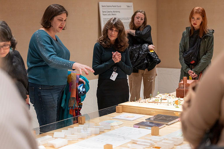 A group of visitors observes a detailed architecture model and sketches displayed on a table, engaging in discussion at a gallery.
