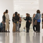 A group of visitors attentively listening to a guide in a bright art gallery, surrounded by modern artwork and sleek white walls.