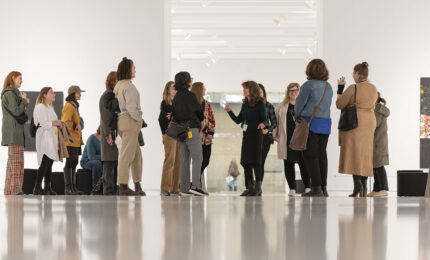 A group of visitors attentively listening to a guide in a bright art gallery, surrounded by modern artwork and sleek white walls.