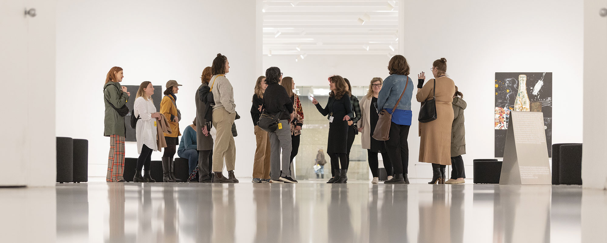 A group of visitors attentively listening to a guide in a bright art gallery, surrounded by modern artwork and sleek white walls.