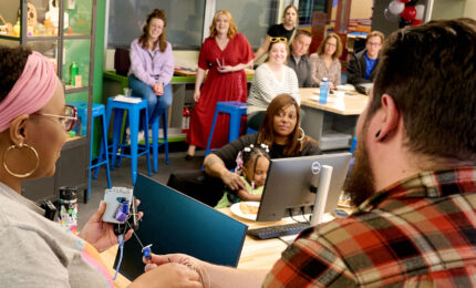 A diverse group of individuals engaged in a learning or collaborative environment, with computers and colorful seating in the background.