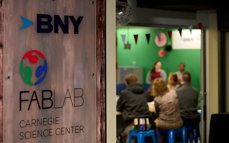 A sign for BNY FABLAB at Carnegie Science Center, with people engaging in activities in a vibrant, decorated workspace in the background.