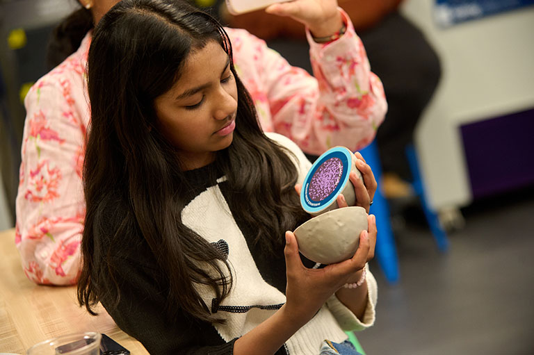 A person holds a small, textured clay bowl and a blue circle tool, engaging in a creative activity in a workshop setting.