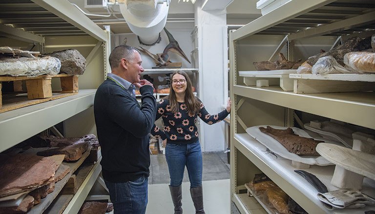 Two individuals explore a storage room filled with shelves of paleontological specimens and fossils.