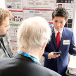 A student presenter in a suit gestures while discussing research with two attendees in front of an academic poster display.