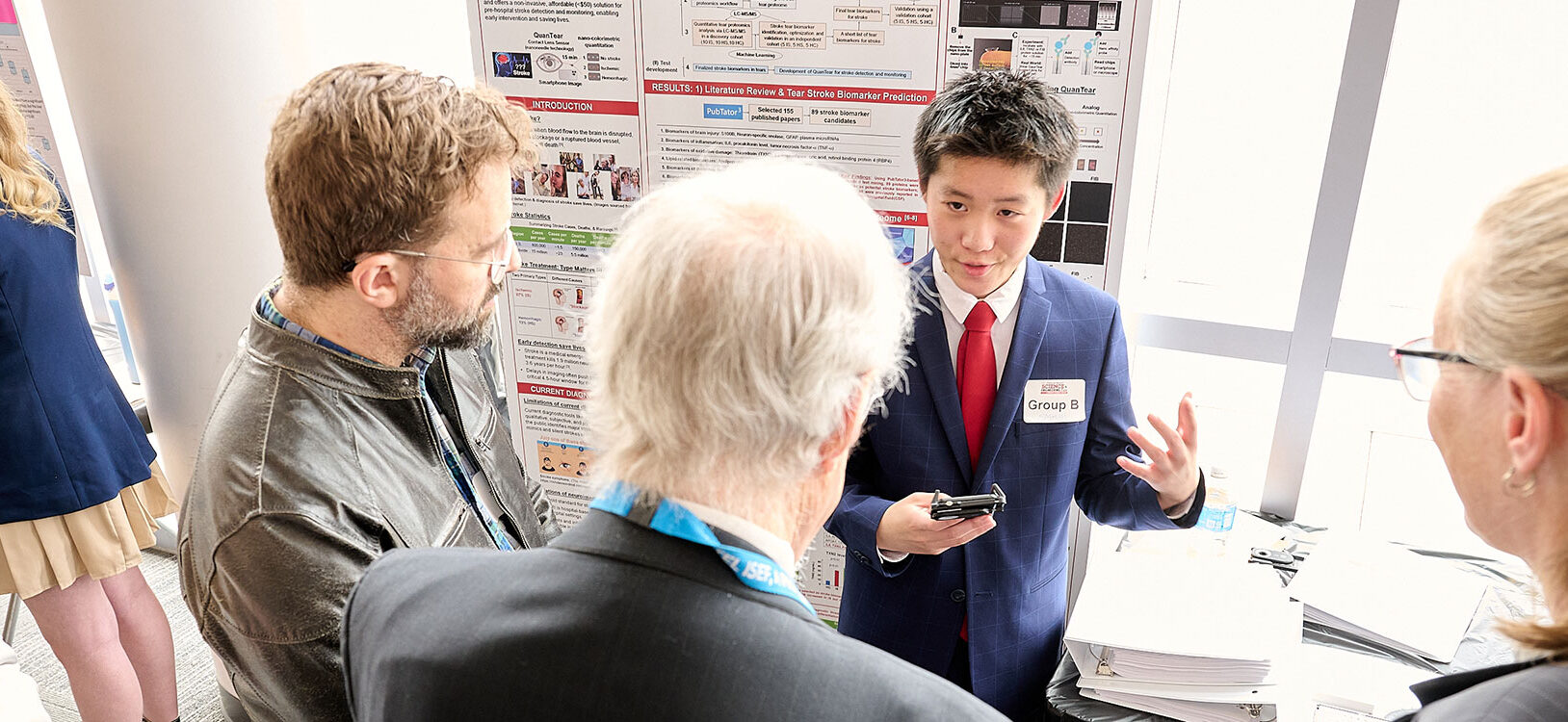 A student presenter in a suit gestures while discussing research with two attendees in front of an academic poster display.
