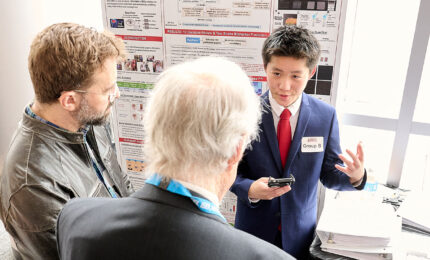 A student presenter in a suit gestures while discussing research with two attendees in front of an academic poster display.
