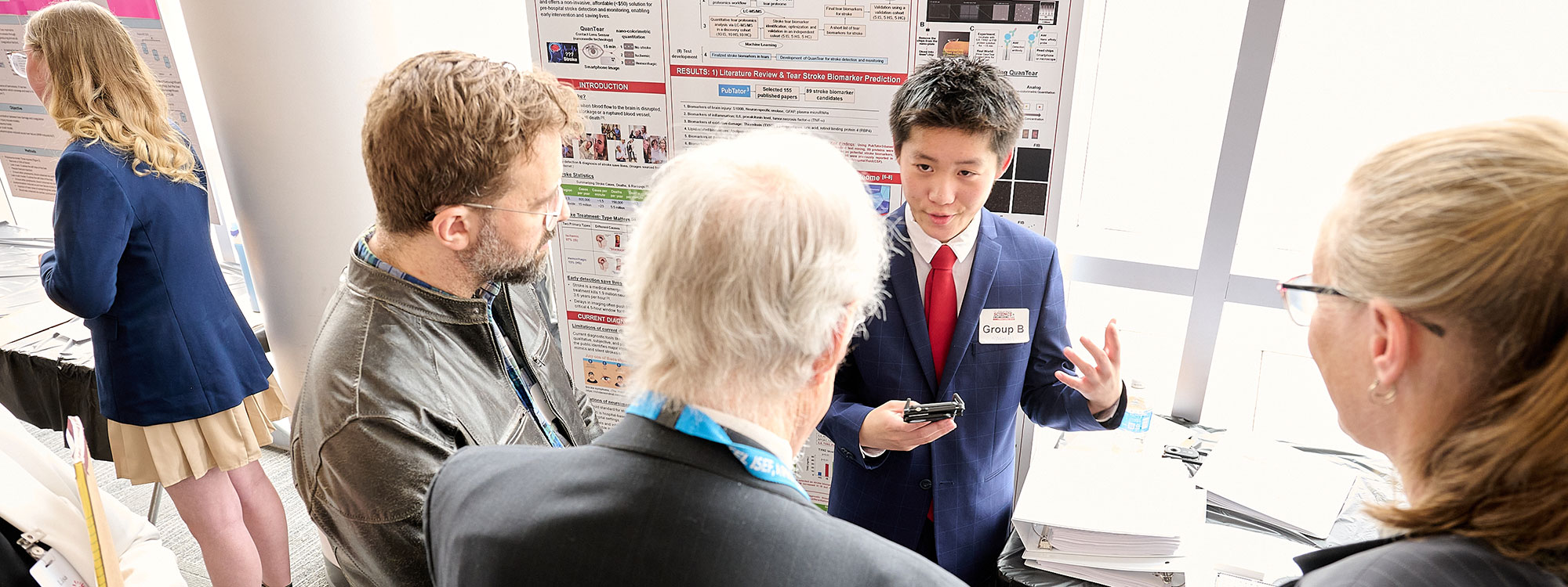 A student presenter in a suit gestures while discussing research with two attendees in front of an academic poster display.