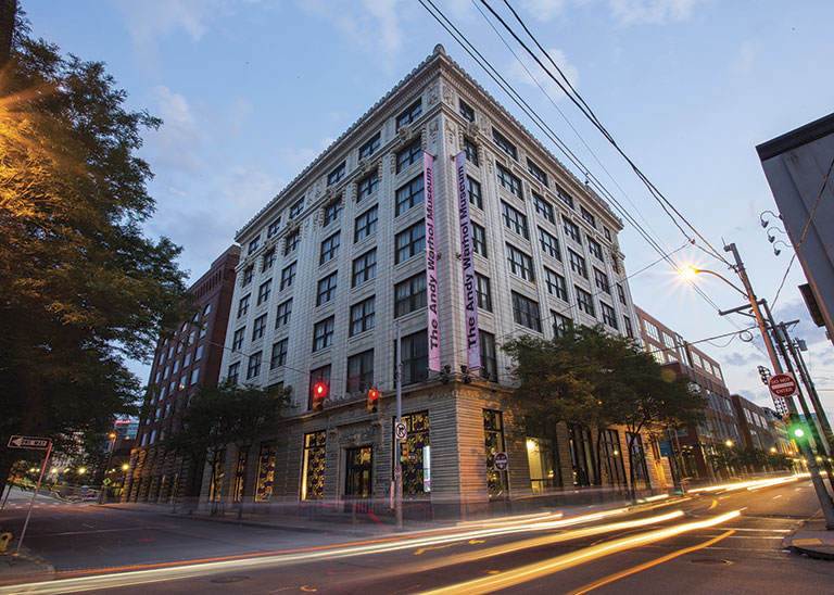 The Andy Warhol Museum, a historic building with eye-catching banners, at dusk, surrounded by city streets and moving traffic.