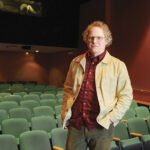 Person wearing a light jacket and red plaid shirt standing alone in a dimly lit empty theater with green seats.