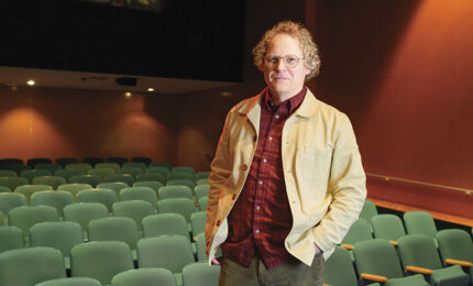 Person wearing a light jacket and red plaid shirt standing alone in a dimly lit empty theater with green seats.