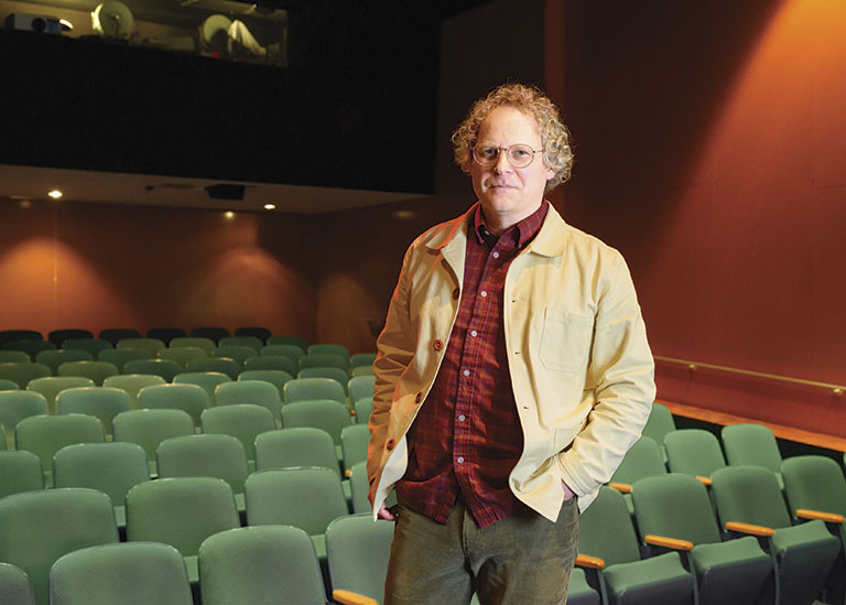Person wearing a light jacket and red plaid shirt standing alone in a dimly lit empty theater with green seats.