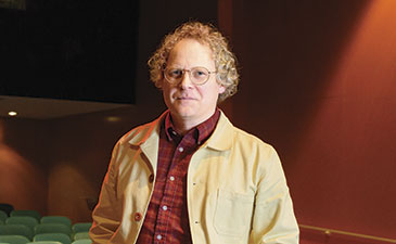 Person wearing a light jacket and red plaid shirt standing alone in a dimly lit empty theater with green seats.