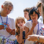 A group of children attentively observe a woman showing them something outdoors. They stand among purple flowers, conveying curiosity and learning.