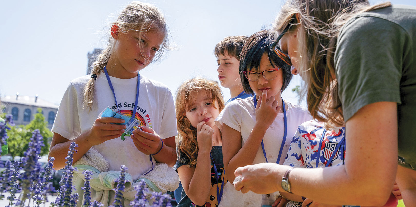 A group of children attentively observe a woman showing them something outdoors. They stand among purple flowers, conveying curiosity and learning.