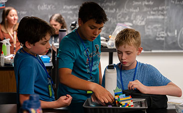 Three boys collaborate on a LEGO project at a classroom table. A chalkboard with writing is in the background, creating a focused learning atmosphere.