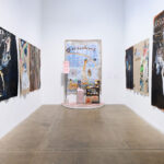 A gallery hallway with colorful, abstract paintings on both walls. The artwork features animals and text. At the end, a small shrine-like installation.