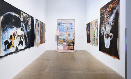 A gallery hallway with colorful, abstract paintings on both walls. The artwork features animals and text. At the end, a small shrine-like installation.