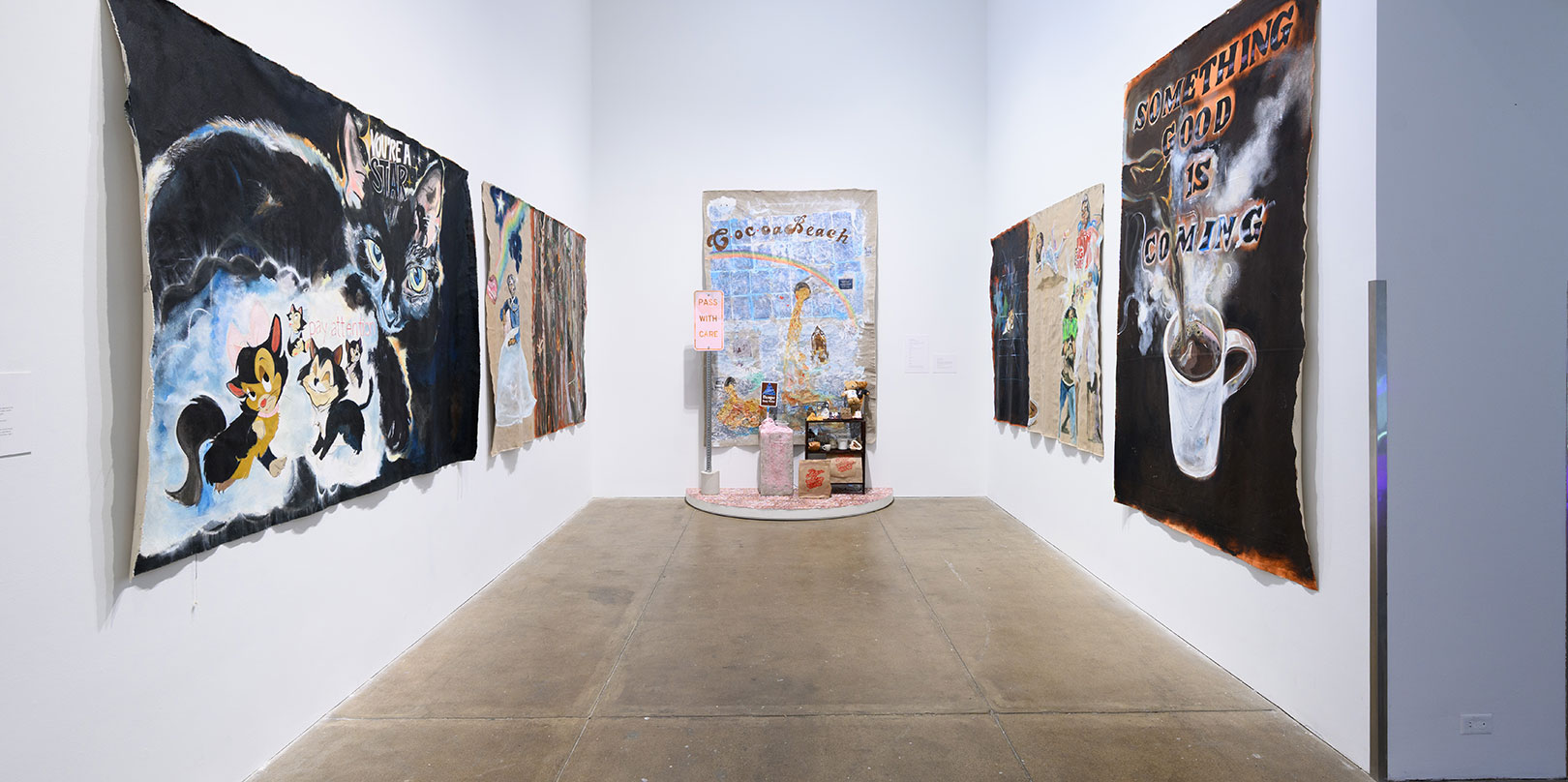 A gallery hallway with colorful, abstract paintings on both walls. The artwork features animals and text. At the end, a small shrine-like installation.