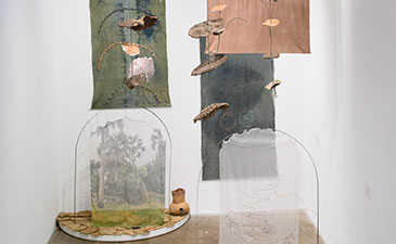 Contemporary art installation features suspended textiles with earthy tones and abstract patterns, organic shapes, and natural textures, creating an introspective ambiance.