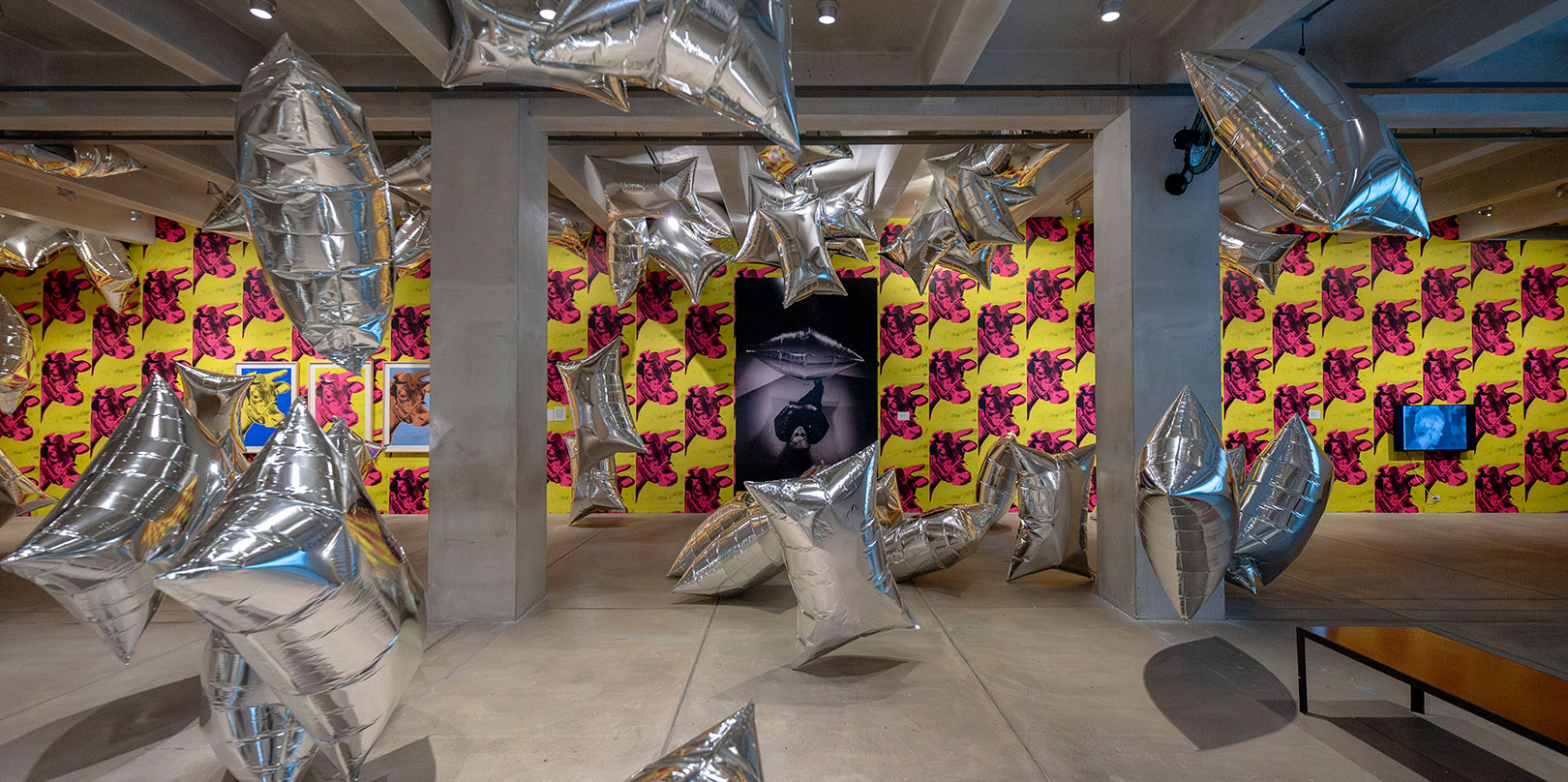 Room filled with floating, silver star-shaped balloons against vibrant yellow and pink wallpaper featuring repeated prints of red cow heads. Bright and whimsical atmosphere.