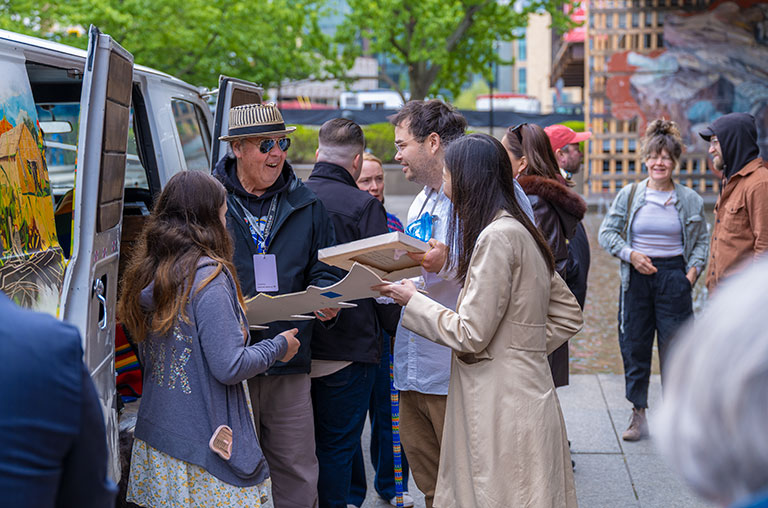A group of people engage in lively conversation near an art van. They hold art pieces, smiling under overcast skies. Urban setting with trees.