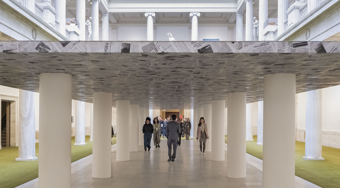 Visitors walk through an art installation resembling a low ceiling supported by white columns inside a classical museum hall.