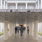 Visitors walk through an art installation resembling a low ceiling supported by white columns inside a classical museum hall.