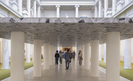 Visitors walk through an art installation resembling a low ceiling supported by white columns inside a classical museum hall.
