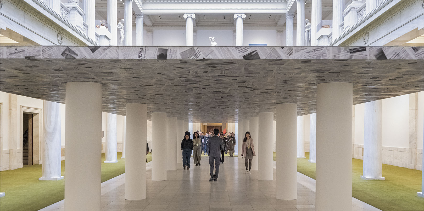 Visitors walk through an art installation resembling a low ceiling supported by white columns inside a classical museum hall.