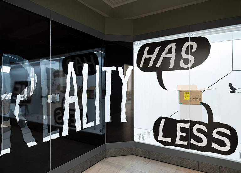 A mirrored display shows the text "REALITY HAS LESS" in bold, distorted letters. The reflective surface creates a fragmented, abstract visual effect.
