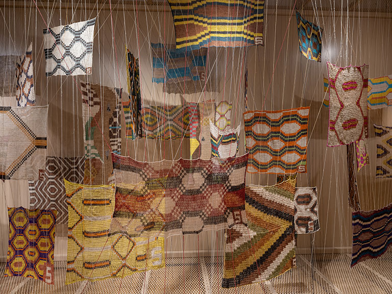 Colorful woven textiles suspended by strings, featuring geometric patterns in various hues and shapes, creating an artistic and dynamic visual display.