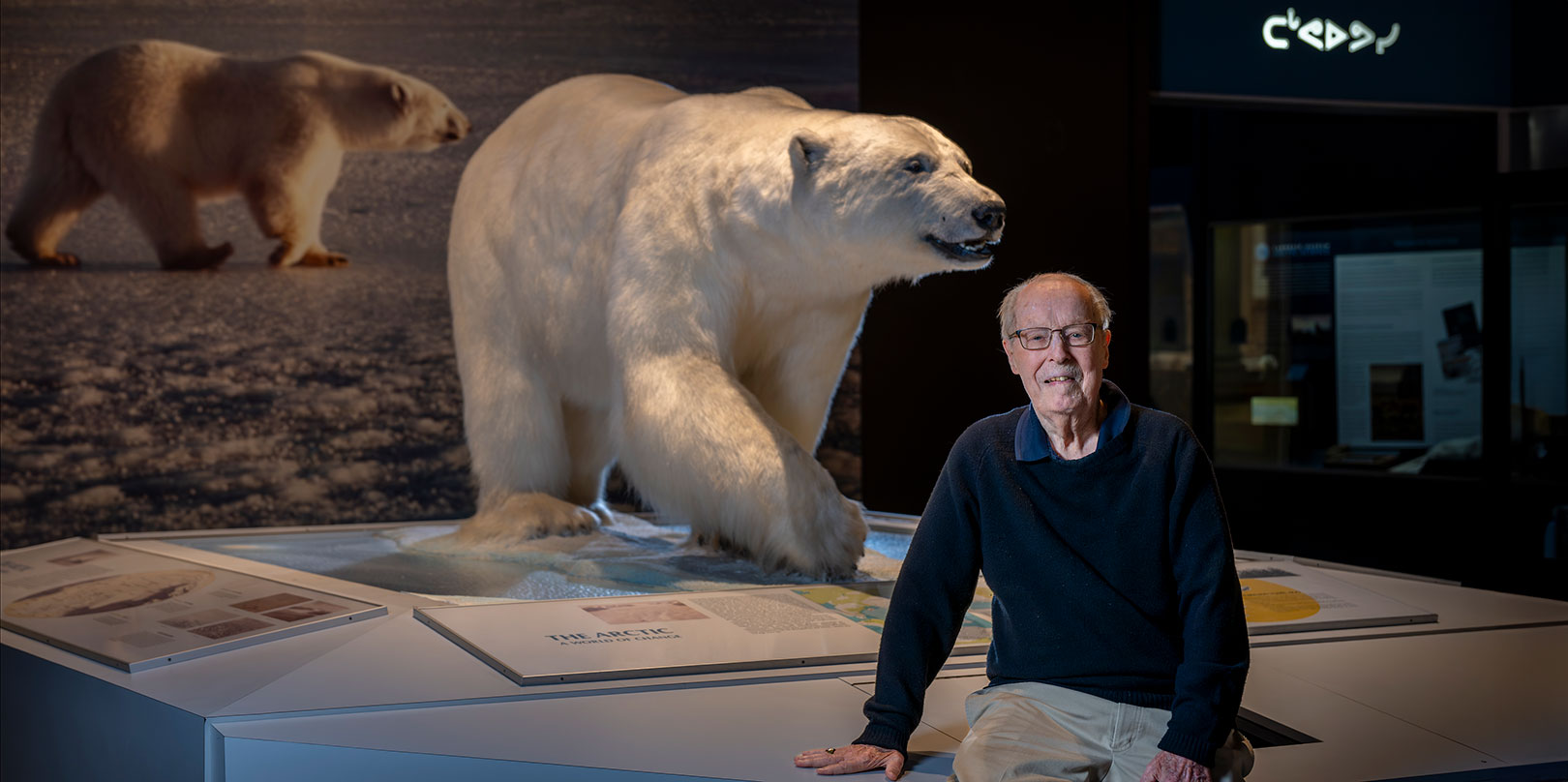 A man sits beside a lifelike polar bear exhibit in a museum. The backdrop features another bear image, creating a harmonious, contemplative atmosphere.
