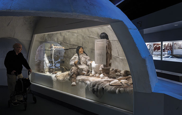 A museum exhibit depicts an Inuit family in an igloo, surrounded by furs and tools. A visitor with a walker observes the scene, creating a historical atmosphere.