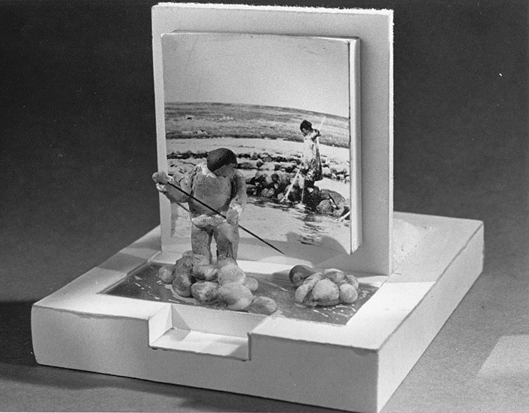 A small diorama features a 3D clay figure spearing fish near rocks in water, set before a black-and-white photo of children doing the same.