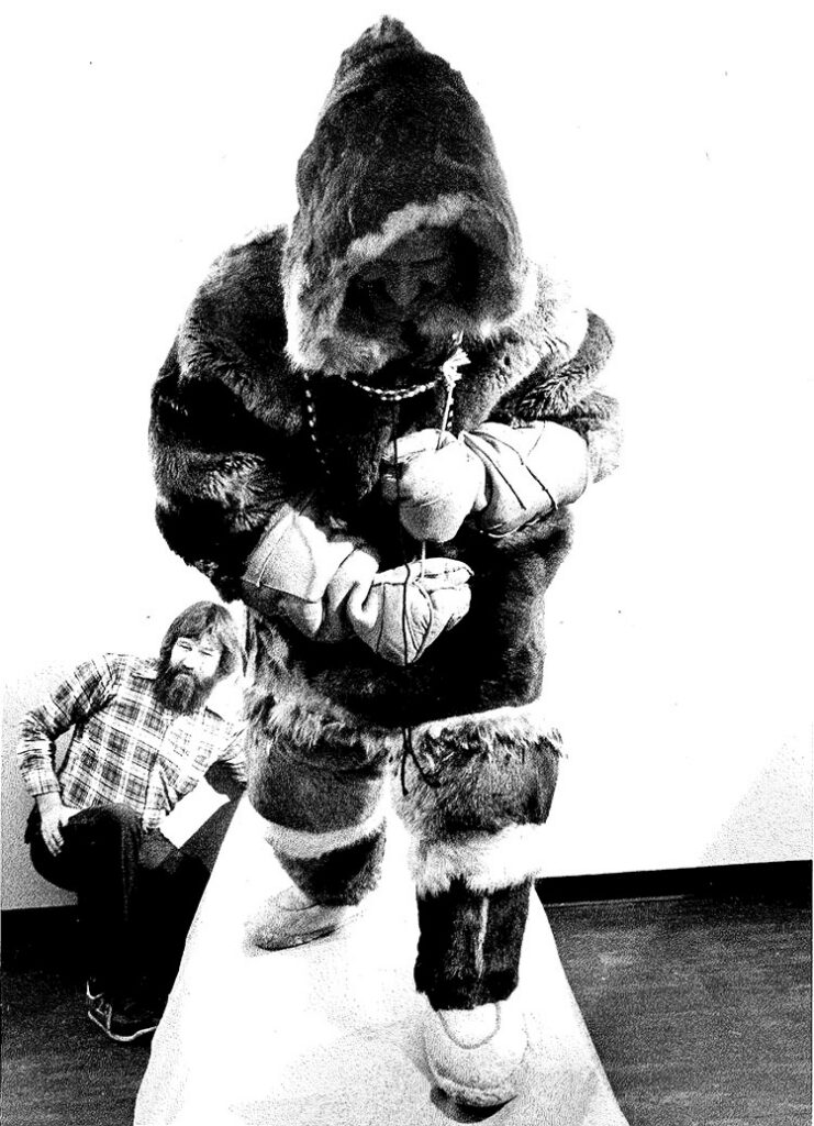 A person in a furry parka and mittens stands poised as if walking. A seated man in a plaid shirt looks on, creating a curious and contemplative scene.