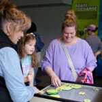 A woman and a child interact with a hands-on educational exhibit. They appear engaged and curious. Informational panels behind them discuss adaptation.