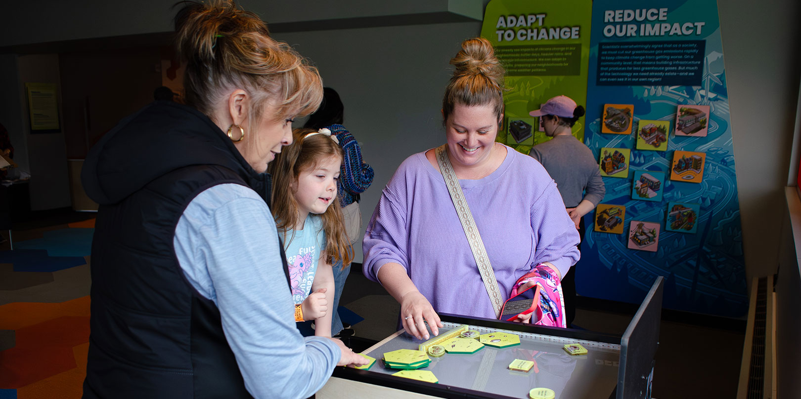 A woman and a child interact with a hands-on educational exhibit. They appear engaged and curious. Informational panels behind them discuss adaptation.