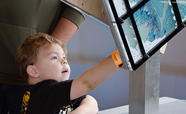 A young child with light brown hair reaches up with curiosity to touch a vertical screen displaying a colorful map, wearing an orange wristband.