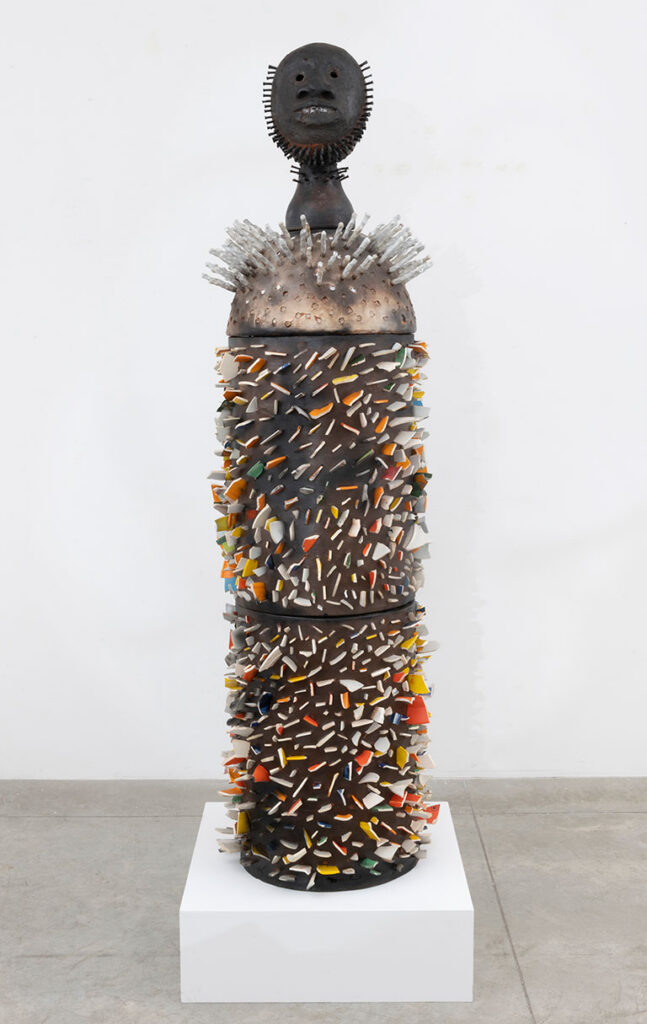 Towering sculpture on a white pedestal, adorned with colorful fragments. It features a spiked, cylindrical body with a meditative, mask-like face.