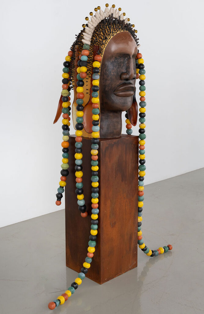 A wooden sculpture of a head in profile with colorful bead strands and a tribal headdress stands on a pedestal against a plain white background.