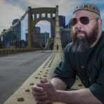 A man wearing sunglasses and a patterned cap leans on a bridge railing, looking thoughtful. The Piitsburgh city skyline and cloudy sky form the backdrop.