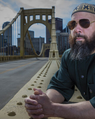 A man wearing sunglasses and a patterned cap leans on a bridge railing, looking thoughtful. The Piitsburgh city skyline and cloudy sky form the backdrop.