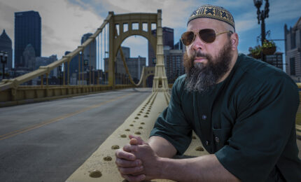 A man wearing sunglasses and a patterned cap leans on a bridge railing, looking thoughtful. The Piitsburgh city skyline and cloudy sky form the backdrop.