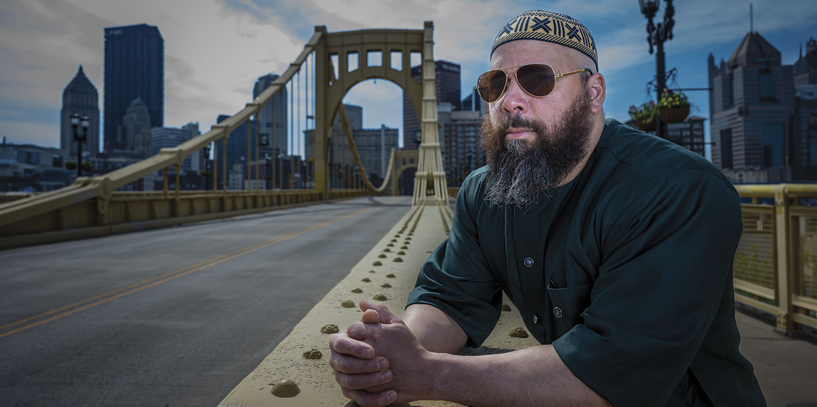 A man wearing sunglasses and a patterned cap leans on a bridge railing, looking thoughtful. The Piitsburgh city skyline and cloudy sky form the backdrop.
