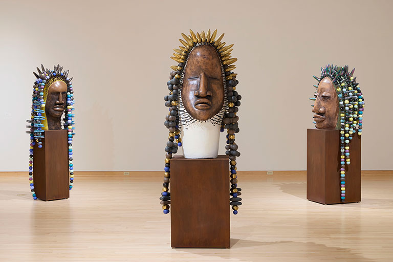 Exhibit features three large wooden masks on pedestals. Each mask has colorful beads or ornaments as hair, set in a bright, minimal gallery.