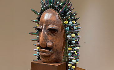 Wooden mask with a serene expression adorned with vibrant spikes and colorful beads. The artwork conveys a sense of cultural richness and creativity.