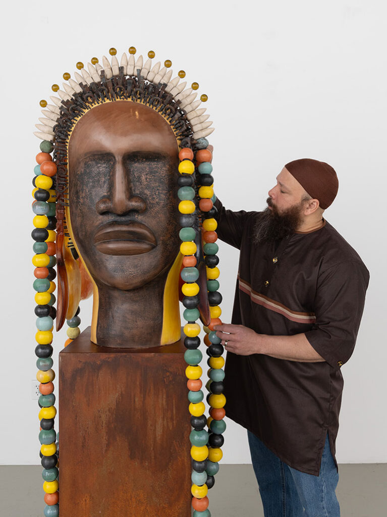 A bearded man adjusts a large, vibrant sculpture of a face with colorful beads and feathers. The setting is a plain room, creating an artistic focus.